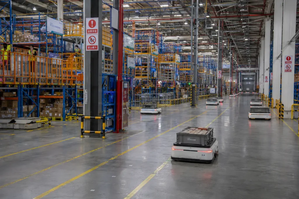 Multiple autonomous mobile robots operating simultaneously on a wide concrete aisle inside a large Chinese e-commerce fulfillment warehouse with tall storage racks and overhead fluorescent lighting